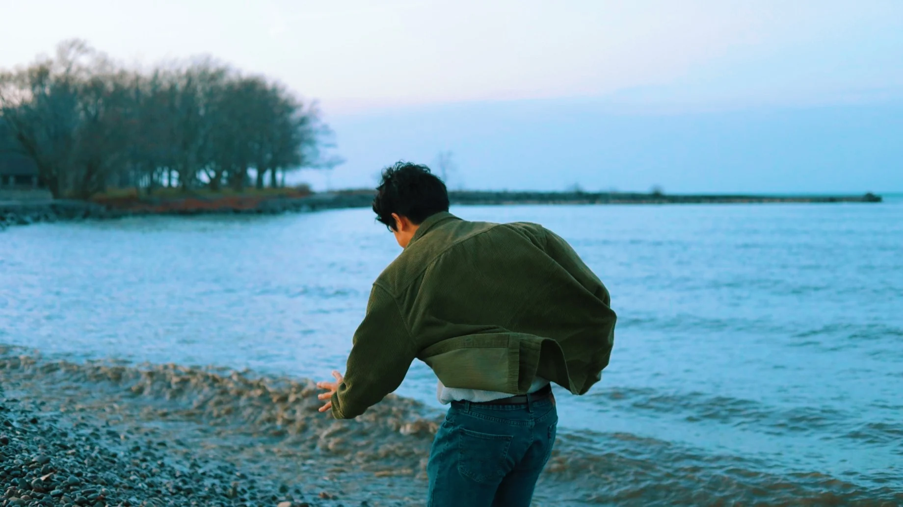 A person leaning toward the water on a pebbled lakeshore