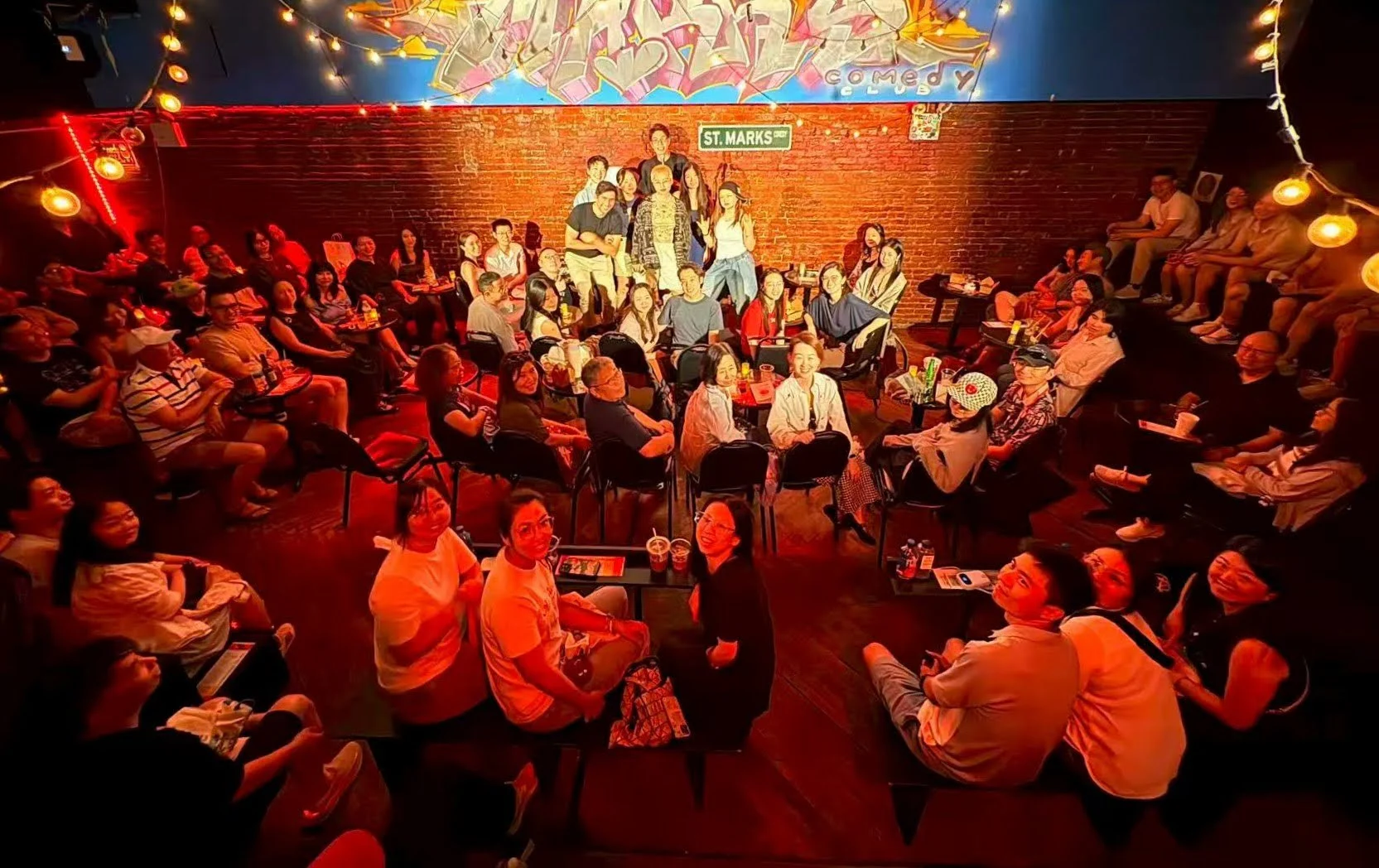 A large group of performers and audience members together in a red-lit comedy venue